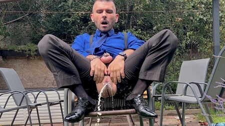 Um homem de fato, com o pénis preso num colete, brinca com o seu ânus e urina em público, vestindo meias transparentes